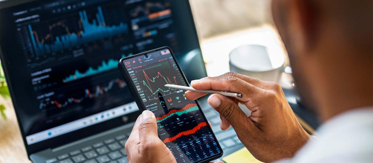 Eine Person prüft Börsenkurse auf einem Smartphone und Laptopbildschirm