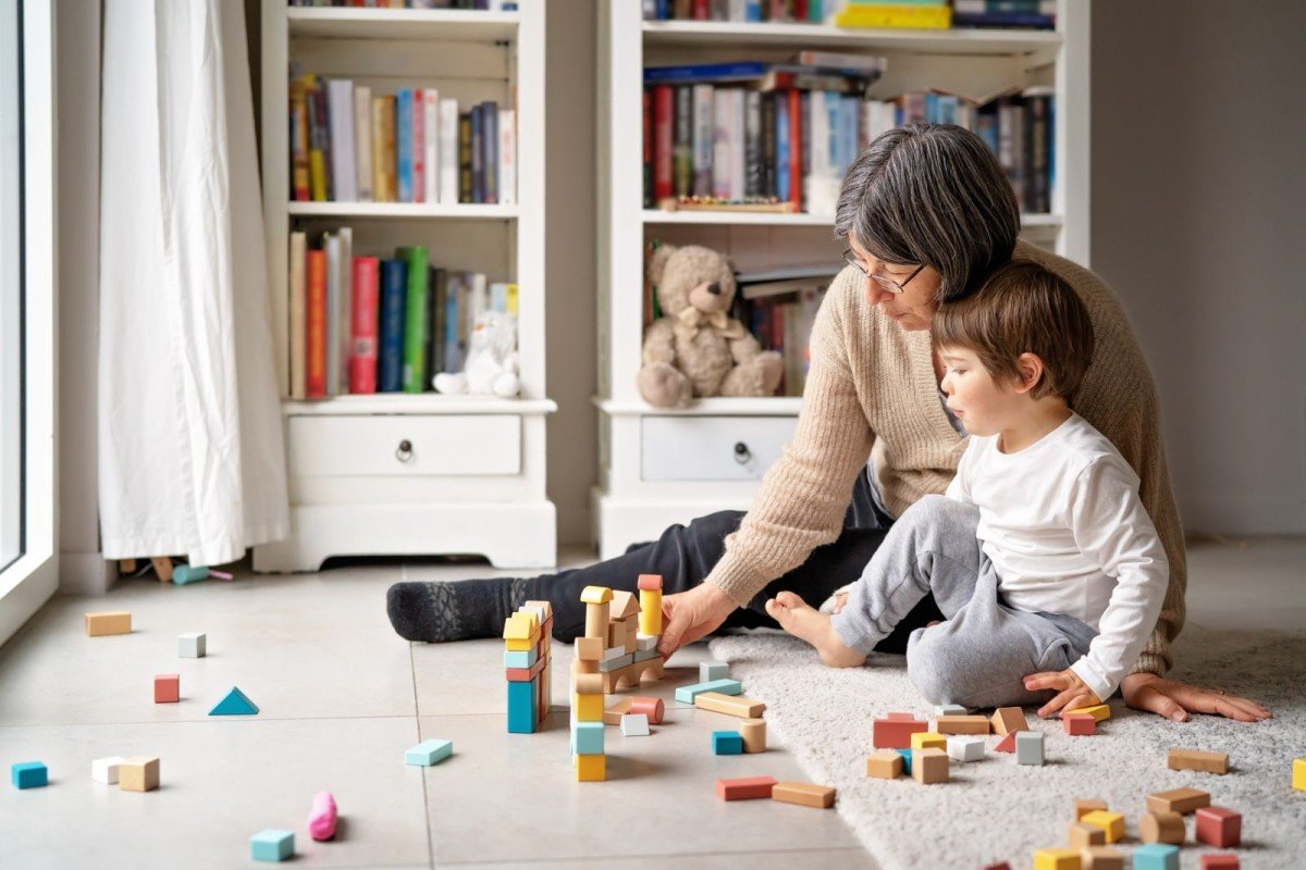 Eine ältere Dame sitzt neben einem Kind am Boden im Wohnzimmer und spielt mit ihm