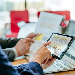 Zwei Personen betrachten Finanzdaten auf Tablet, Laptop und Papier.