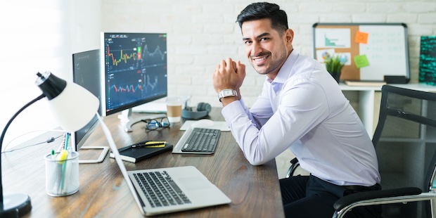 A man sits at a desk before multiple monitors with stock charts, smiling - neutral trading motif