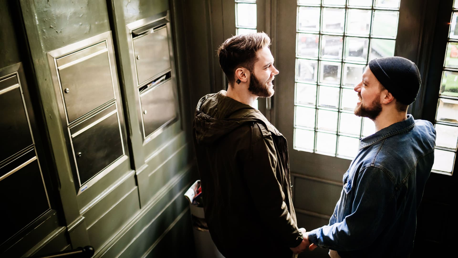 Zwei Männer, die sich an den Händen halten und lächelnd angucken stehen in einem Hausflur vor der Haustür.