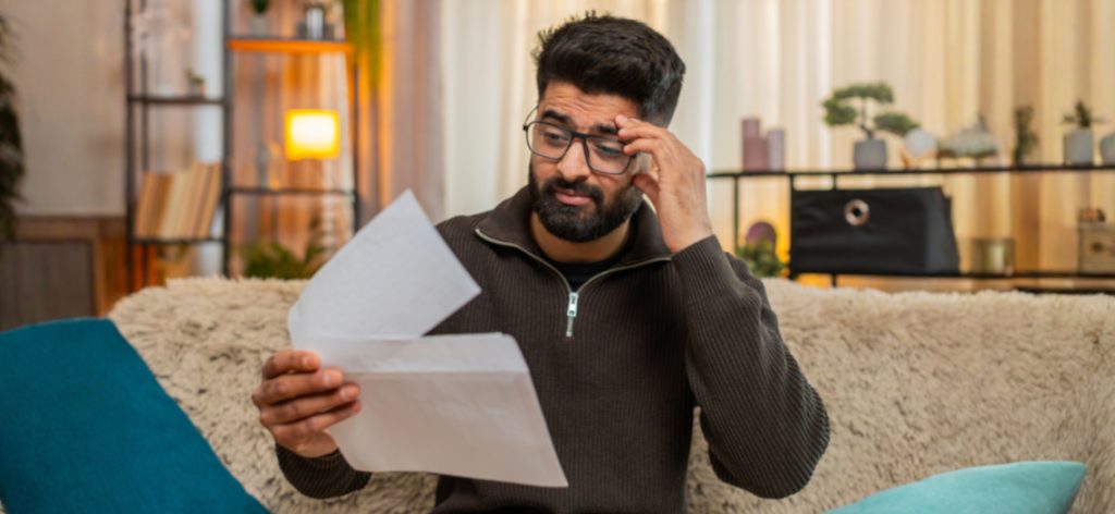 A man with a beard and glasses sitting on a sofa in a living room and reading documents