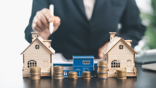 Person using stylus behind miniature houses with stacked coins on desk